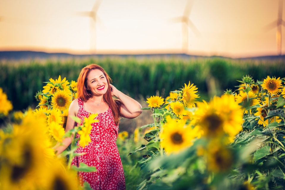 A smiling woman in a patterned red dress appears among blooming sunflowers.