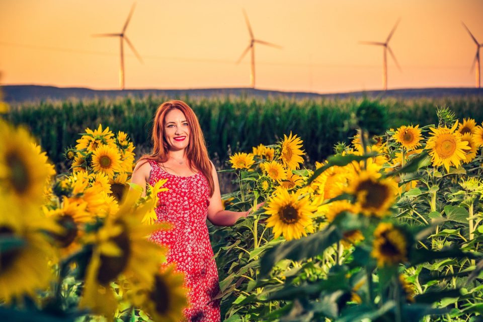 A smiling woman in a patterned red dress poses among blooming sunflowers.