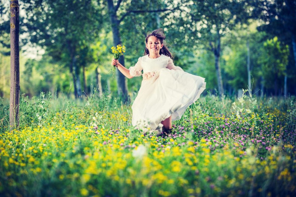 Una niña con vestido blanco de comunión corre sonriente por un campo cubierto de flores silvestres mientras sostiene.