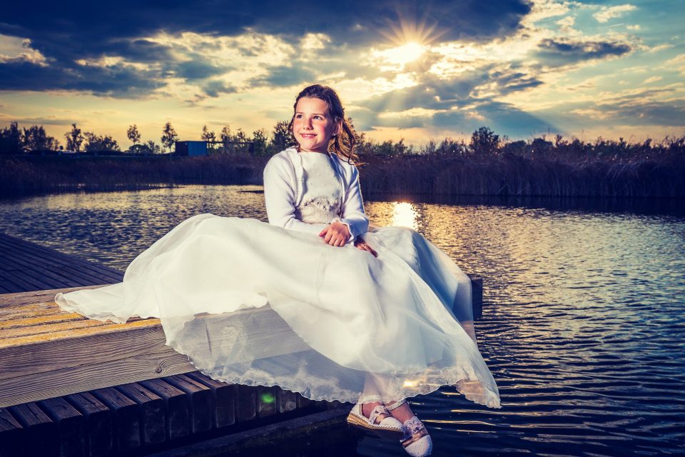 Una niña con vestido blanco de comunión está sentada en un embarcadero de madera junto a un estanque.