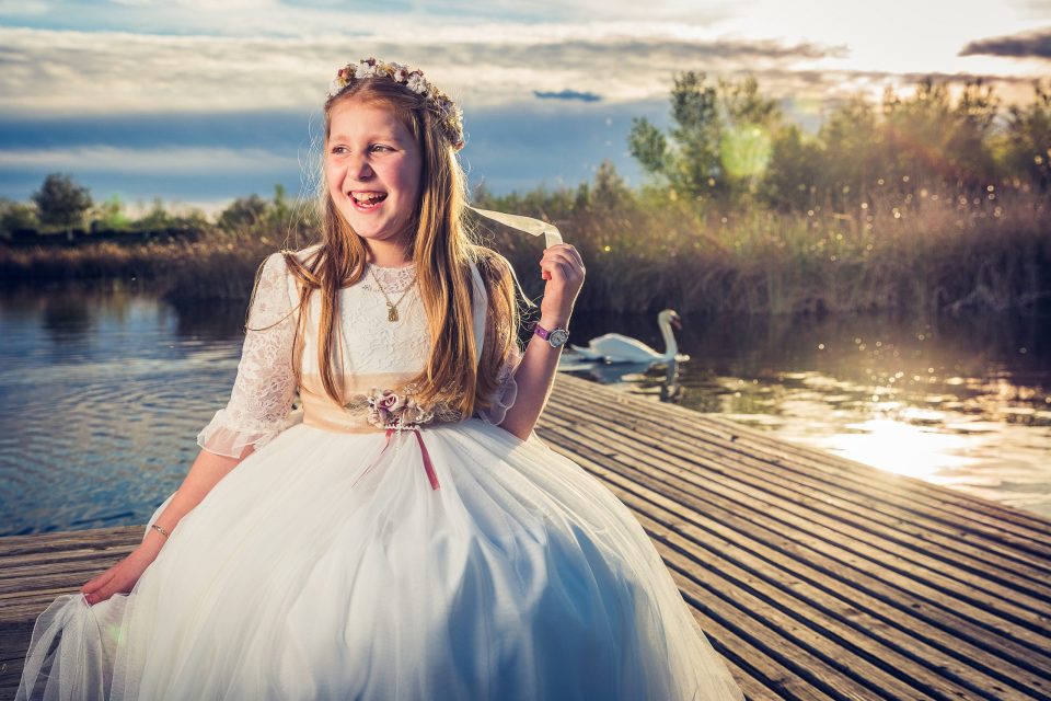 Una niña con vestido blanco de comunión y corona de flores sonríe sentada en un muelle de madera junto al agua.