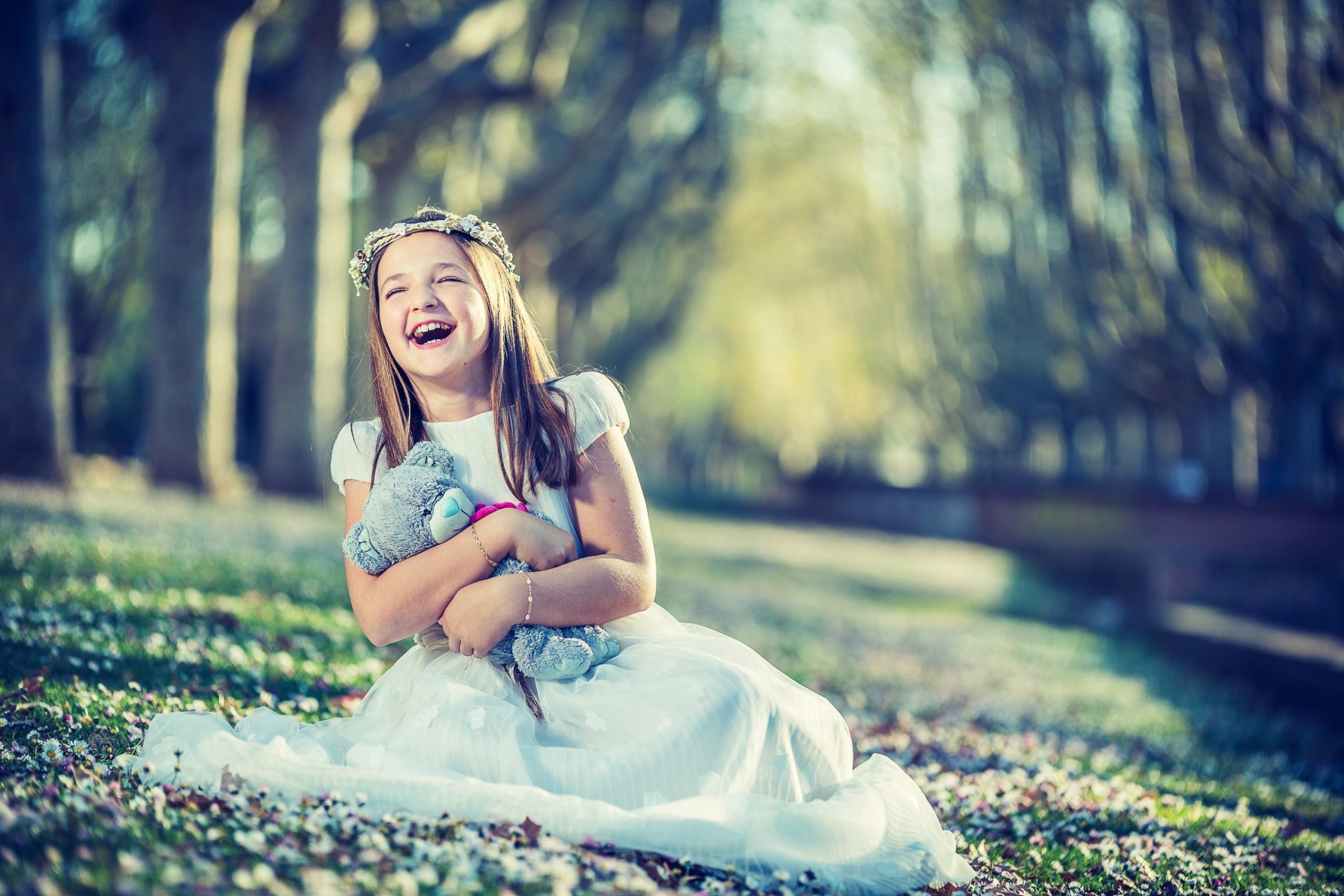 Una niña con vestido blanco y una corona floral está sentada sobre la hierba de un parque.