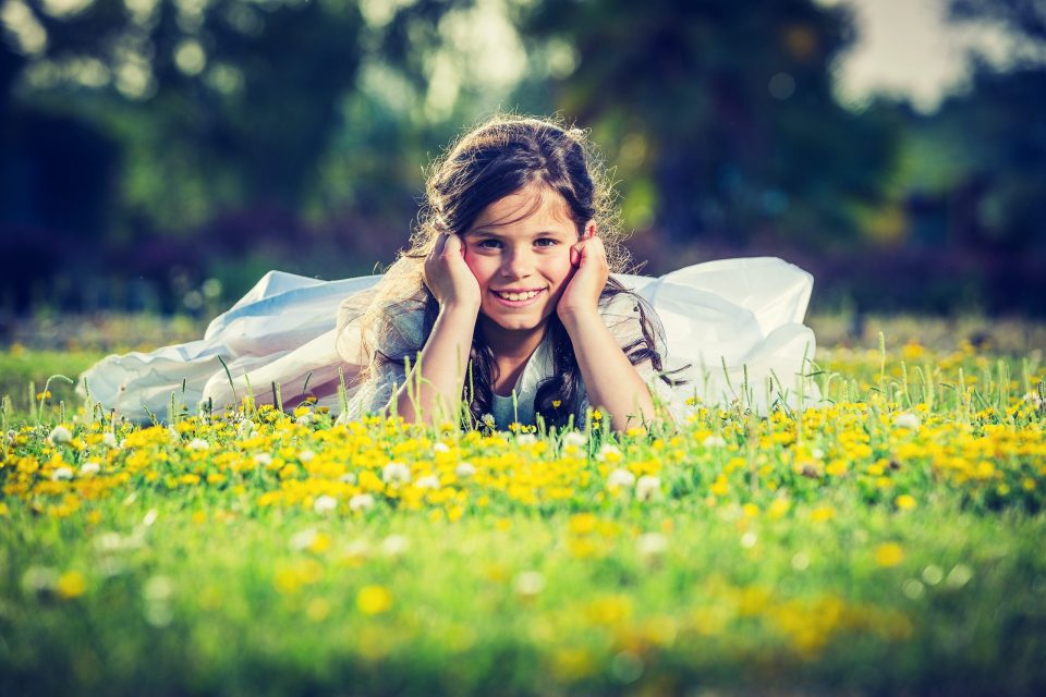 Una niña sonríe tumbada boca abajo entre flores amarillas y blancas en un campo.