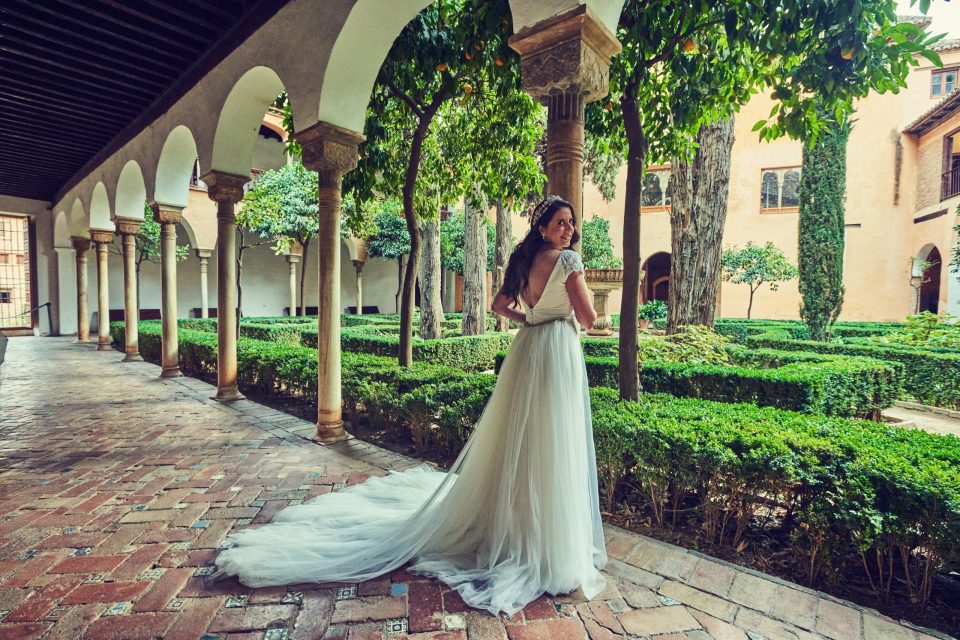 Una novia con vestido blanco de cola larga posa y mira hacia atrás en un patio ajardinado.