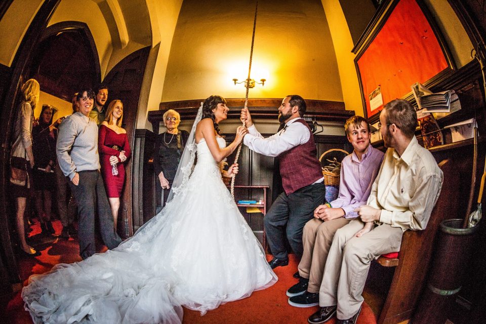 A bride in a white dress and veil and a groom in a dark waistcoat sway together on a rope swing indoors.