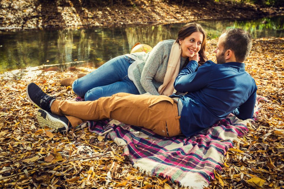 An adult couple relaxes on a checked blanket among fallen leaves beside a tree-lined pond.