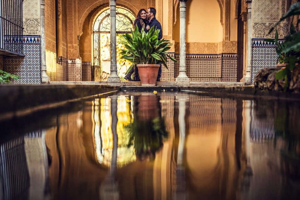 A couple embraces beneath an ornate arch in an interior courtyard decorated with geometric tiles and columns.