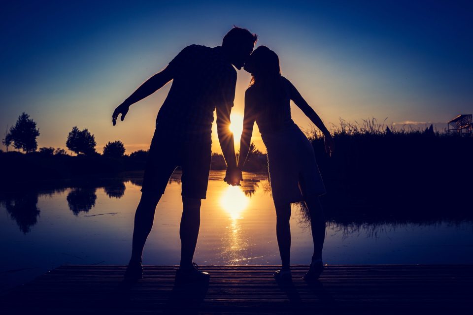 Una pareja aparece en silueta sobre un embarcadero de madera, besándose y sujetándose de la mano frente a un estanque.