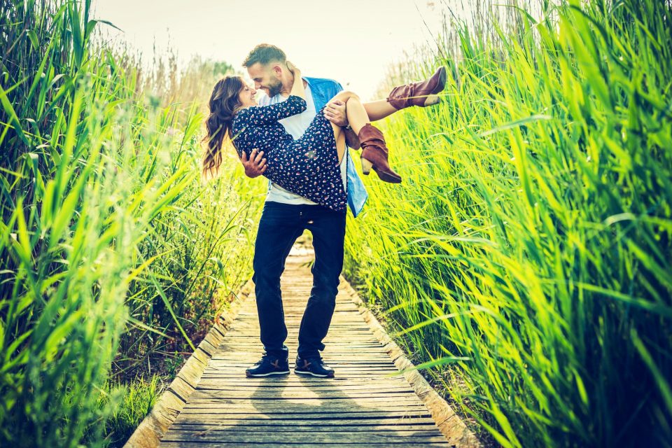 Una pareja aparece en un sendero de madera rodeado de hierbas altas y vegetación abundante.