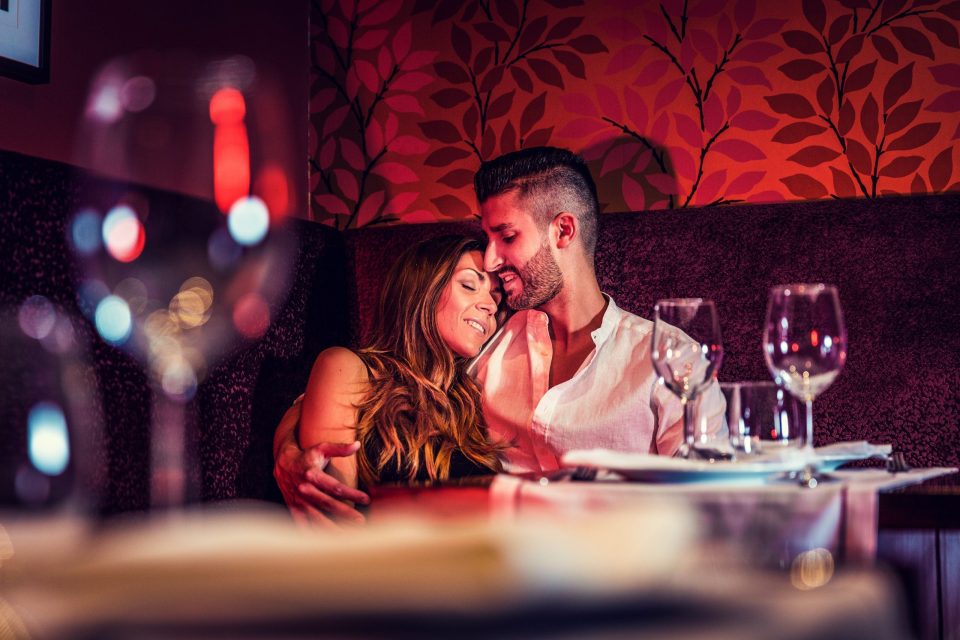 Una pareja aparece sentada en un comedor de hotel, abrazada y sonriendo con los ojos cerrados.