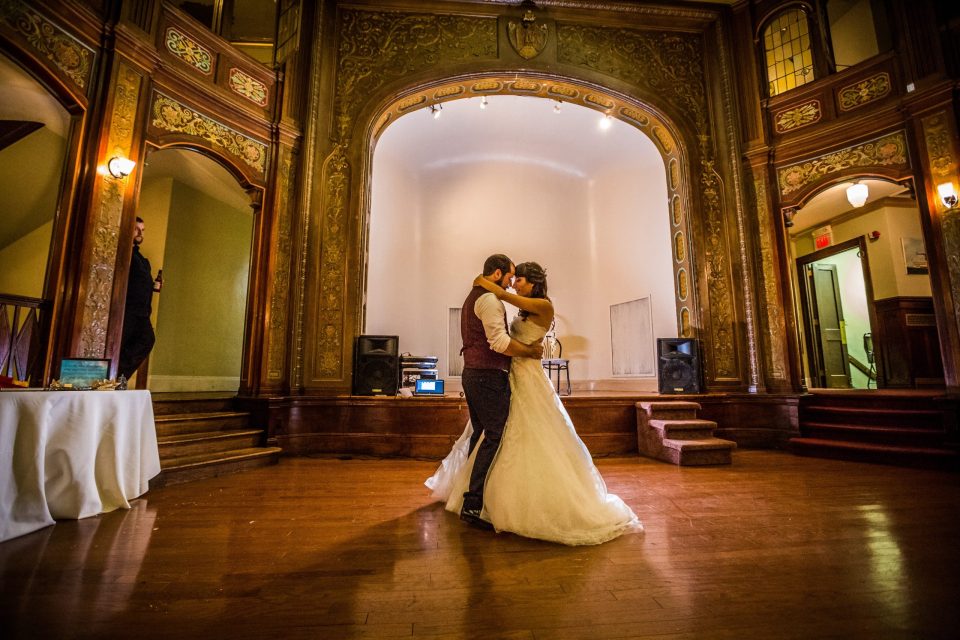 A couple dance embracing in the centre of an ornate hall with gilded mouldings.