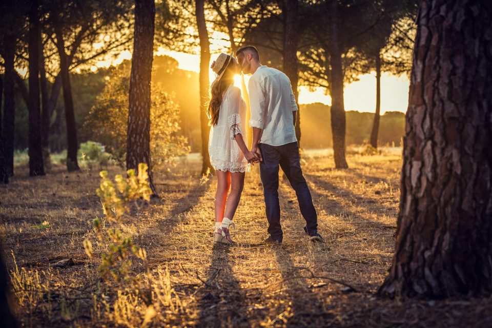 Una pareja camina de la mano entre pinos mientras se acerca y se besa.
