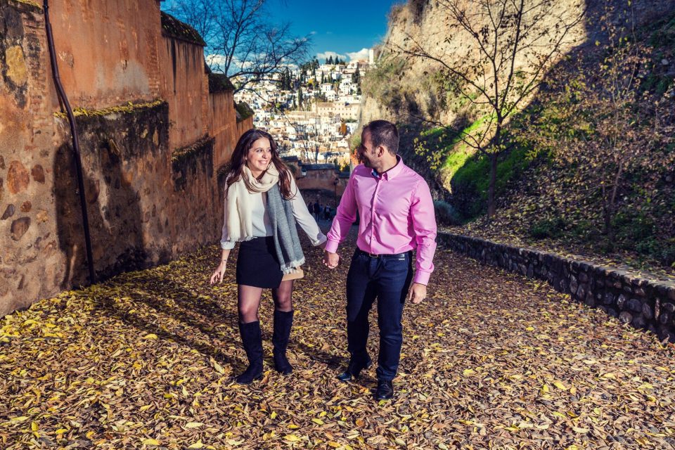 A couple walk hand in hand along a path covered with dry leaves, between ancient stone walls and vegetation.