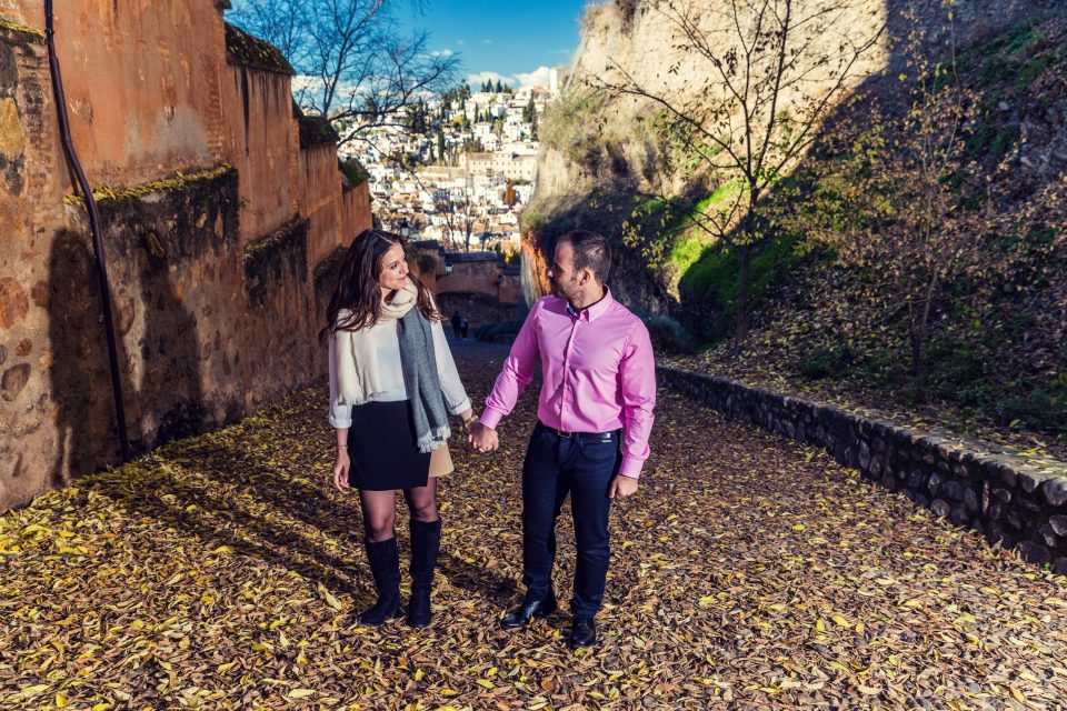 Una pareja camina de la mano por un sendero exterior cubierto de hojas, entre muros antiguos y vegetación.