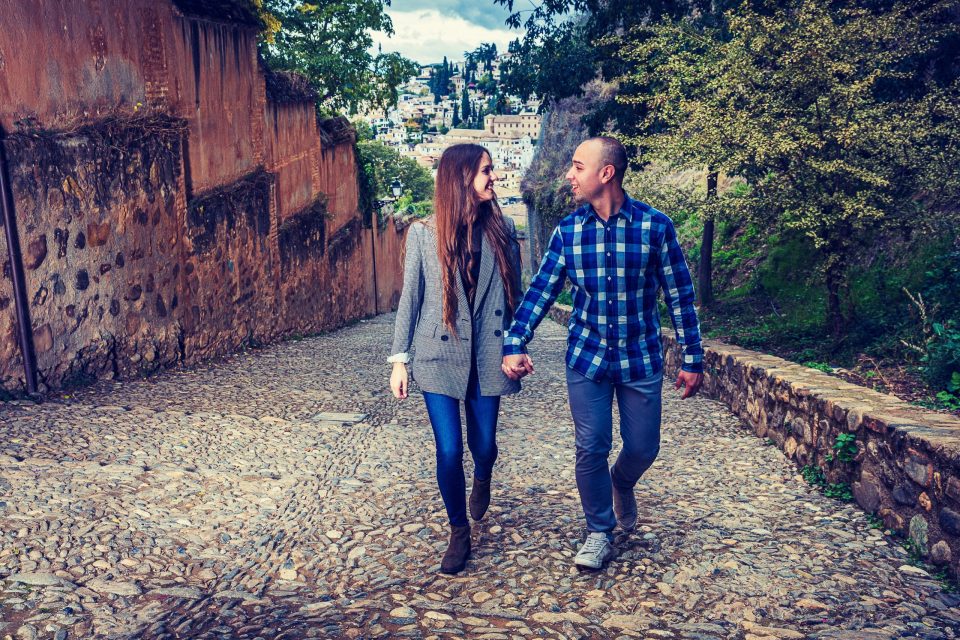 Una pareja camina de la mano por una calle empedrada entre antiguas murallas de piedra.