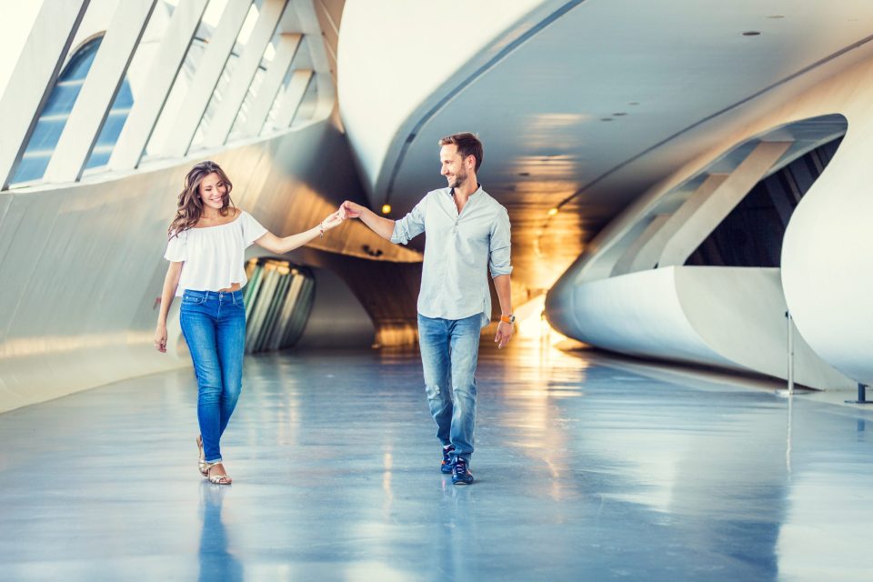 Una pareja camina de la mano y sonríe en el interior de un espacio moderno y amplio, rodeado de grandes ventanales.