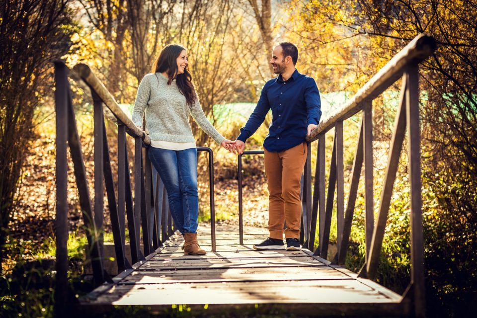 A couple walk hand in hand and smile on a wooden bridge surrounded by trees with golden leaves.