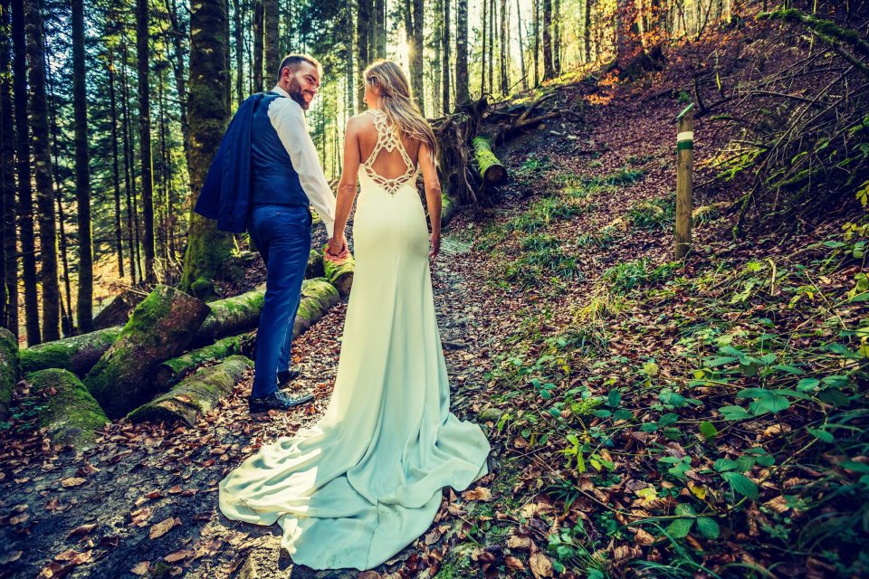 Una pareja con atuendo nupcial camina de la mano por un sendero de montaña rodeado de un bosque frondoso.