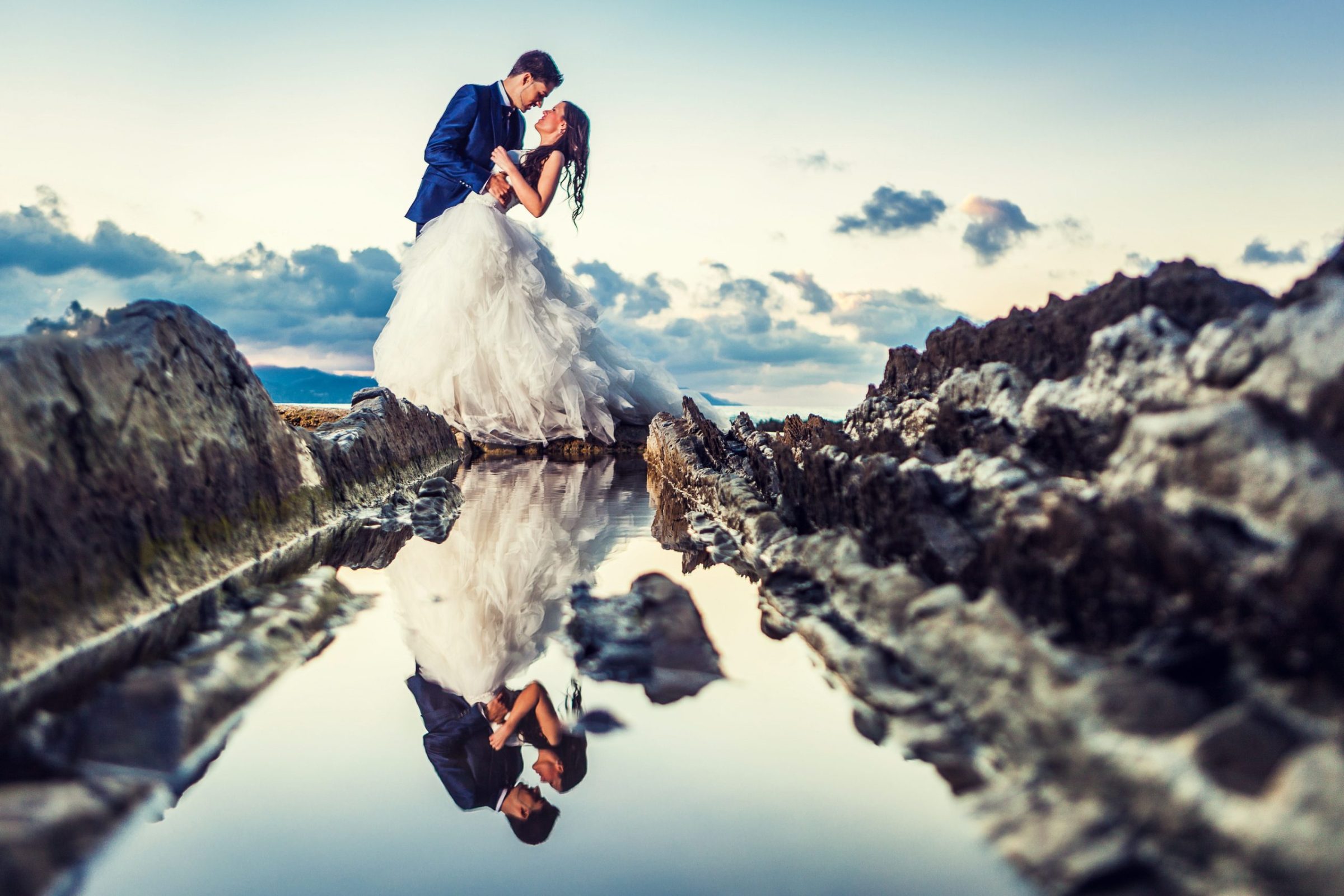 A couple in wedding attire pose embracing on a rocky shore beside a pool of water.