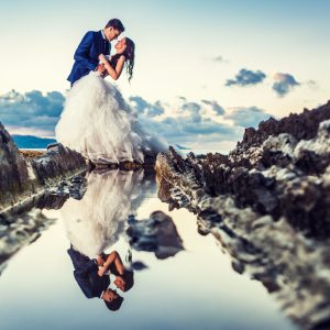 A couple in wedding attire pose embracing on a rocky shore beside a pool of water.
