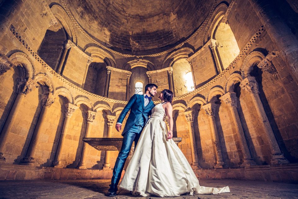 A couple in wedding attire pose inside a church or historic stone building.
