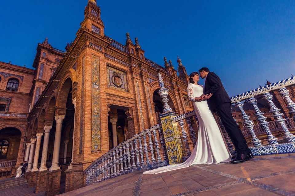 A couple in wedding attire poses on a staircase decorated with railings and ornamental tiles.