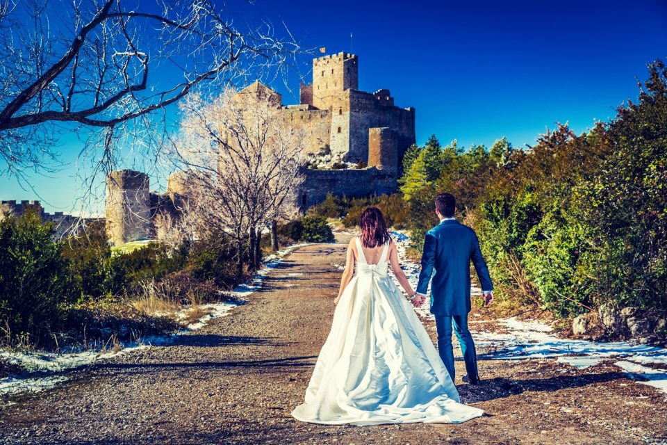 Una pareja de novios camina de espaldas por un sendero, de la mano, frente a un castillo medieval.