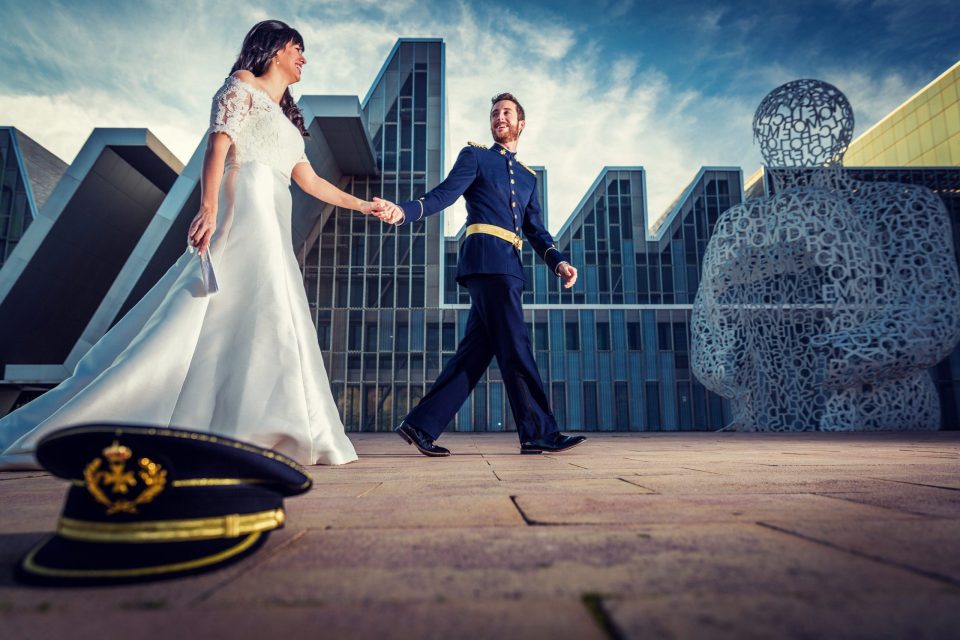 A newlywed couple walk hand in hand in front of a modern building.