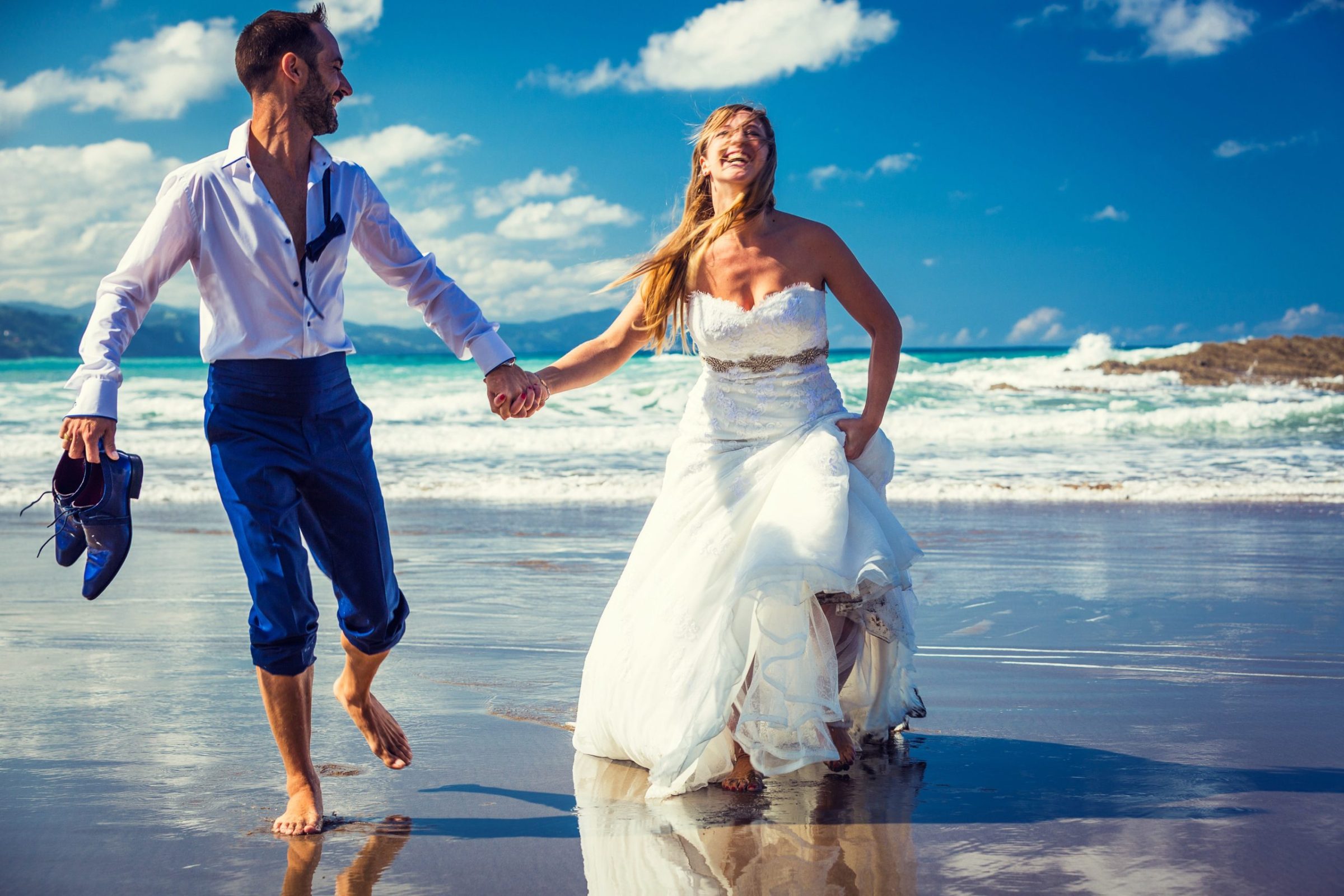 A newlywed couple walks barefoot along the seashore hand in hand.