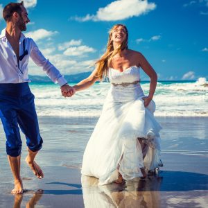 A newlywed couple walks barefoot along the seashore hand in hand.