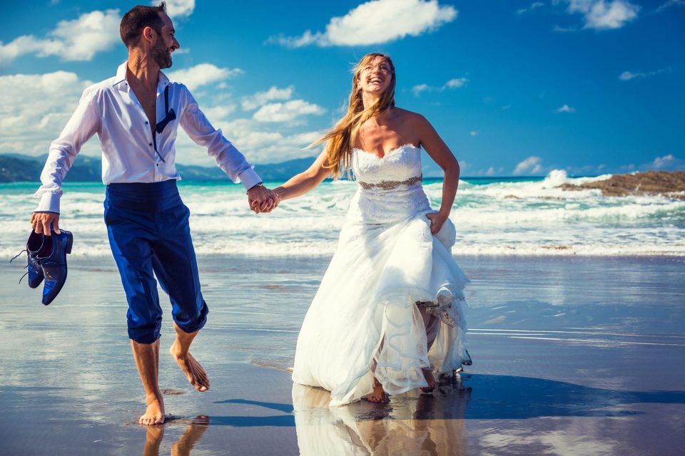 A newlywed couple walks barefoot along the seashore hand in hand.