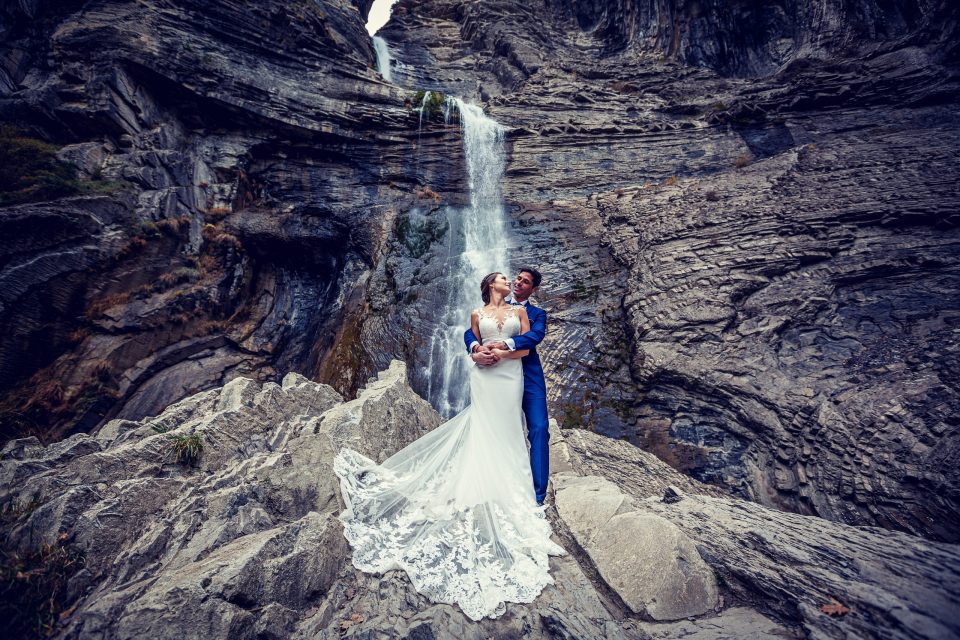 Una pareja de novios posa abrazada sobre unas rocas frente a una cascada, en un paisaje montañoso de paredes de roca.