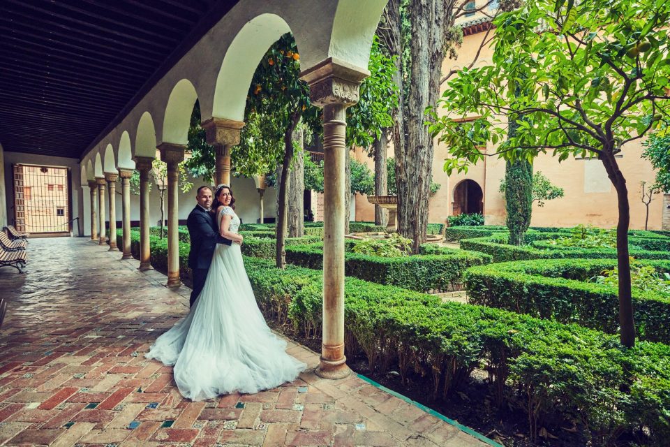 Una pareja de novios posa bajo una galería de arcos en un patio ajardinado de un monumento.