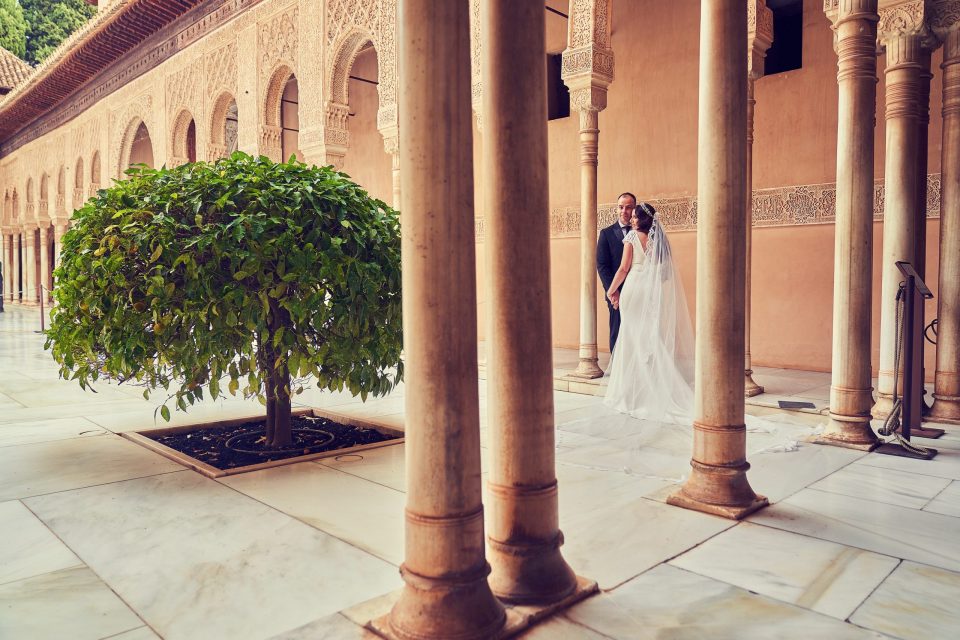 Una pareja de novios posa bajo una galería de columnas, rodeada de arcos decorados con relieves ornamentales.