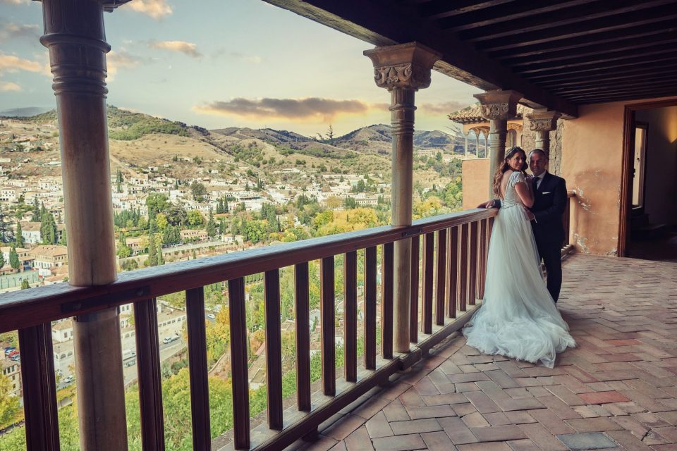 Una pareja de novios posa en el balcón porticado de un edificio monumental, rodeada de columnas de piedra y vigas de madera.