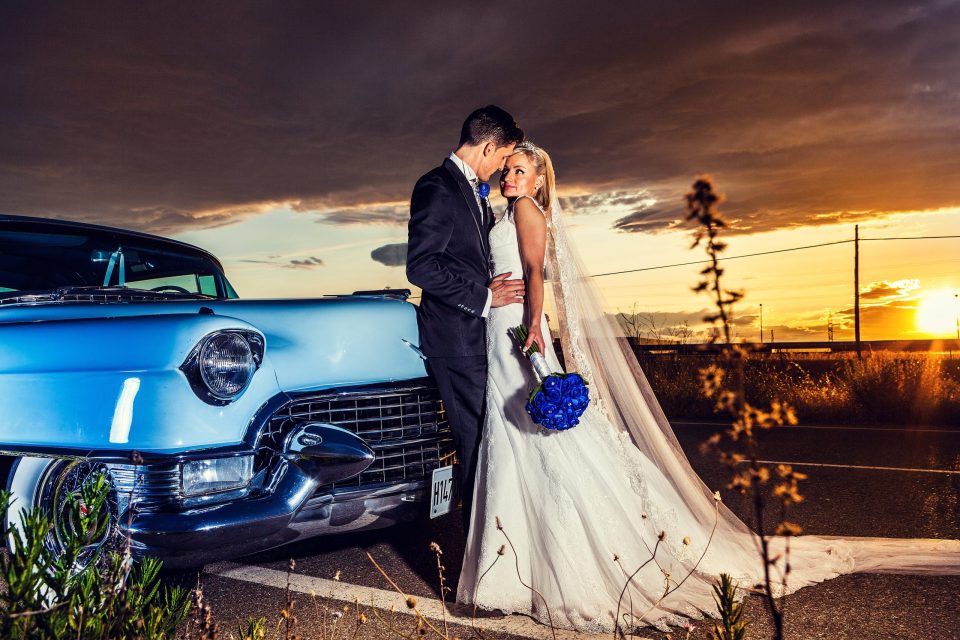Una pareja de novios posa junto a un automóvil clásico azul al atardecer.