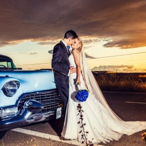 A newlywed couple poses beside a blue classic car at sunset.