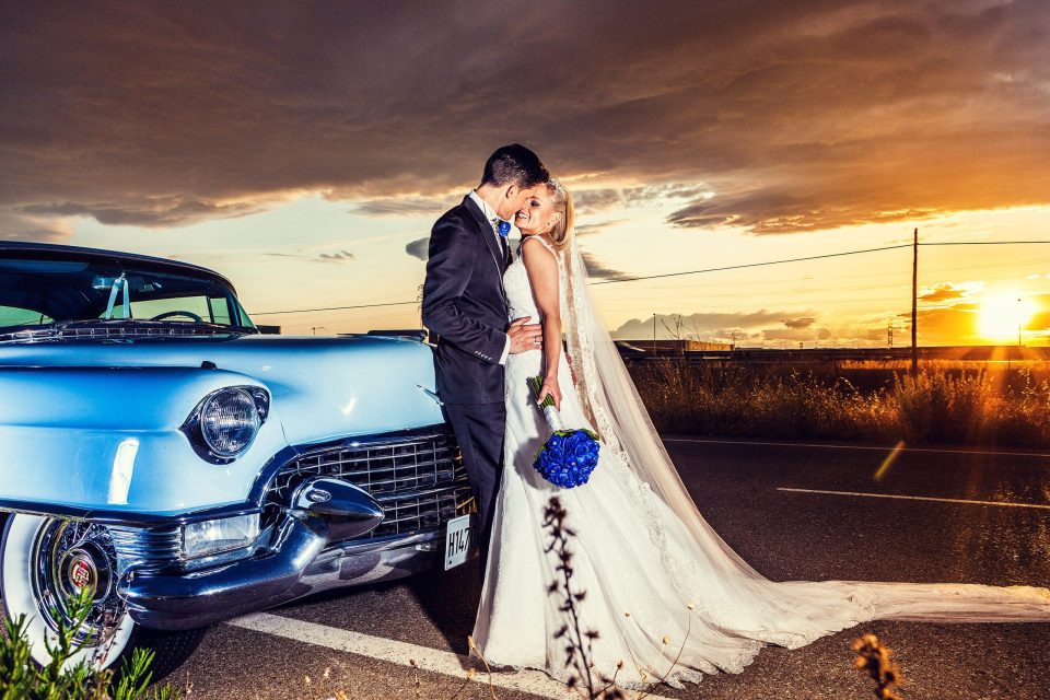 A newlywed couple poses beside a blue classic car at sunset.