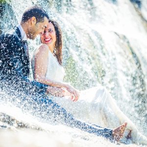 A bride and groom sit beside a waterfall or stream, surrounded by rocks and vegetation.