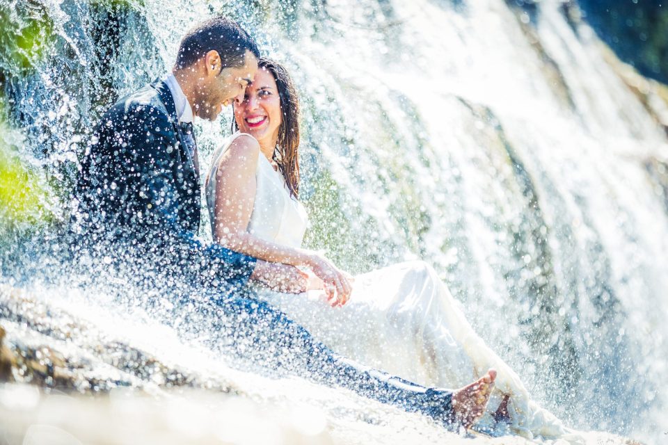 Una pareja de novios posa sentada junto a una cascada o corriente de agua, rodeada de rocas y vegetación.