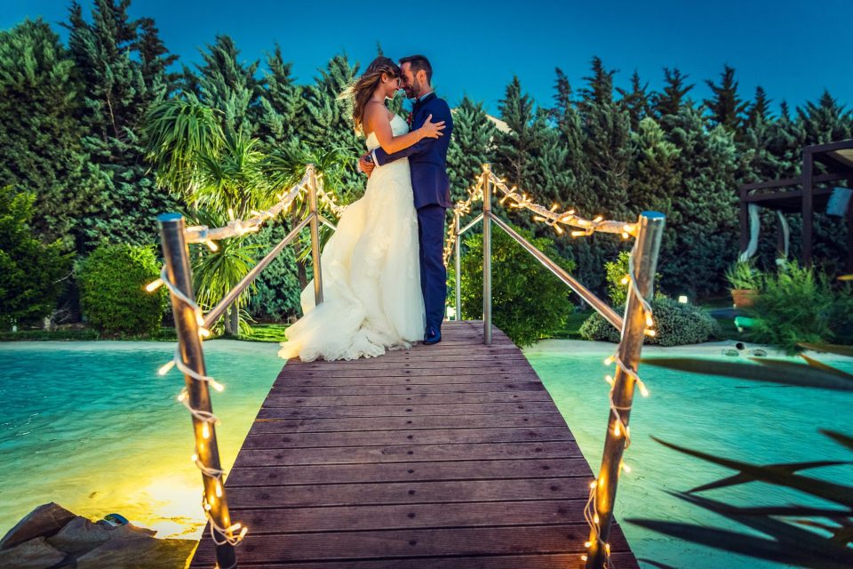 Una pareja de novios se abraza sobre un puente de madera que cruza una piscina en el exterior de una finca.