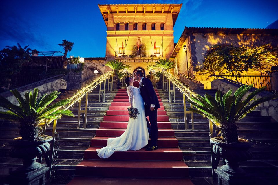 Una pareja de novios se besa en el centro de unas escaleras con alfombra roja frente a una finca iluminada durante la noche.