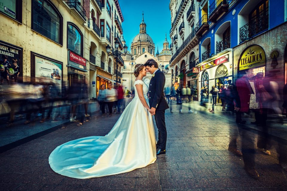 Una pareja de novios se besa en el centro de una calle peatonal urbana, rodeada de edificios históricos.
