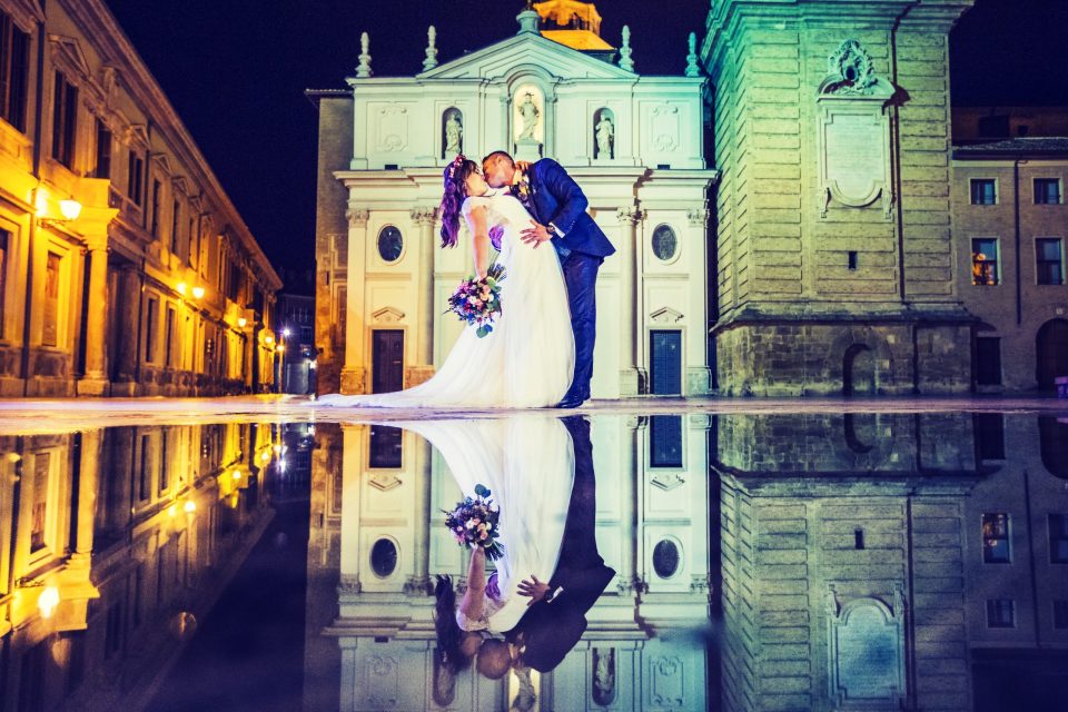 Una pareja de novios se besa frente a una iglesia iluminada, en una plaza urbana durante la noche.