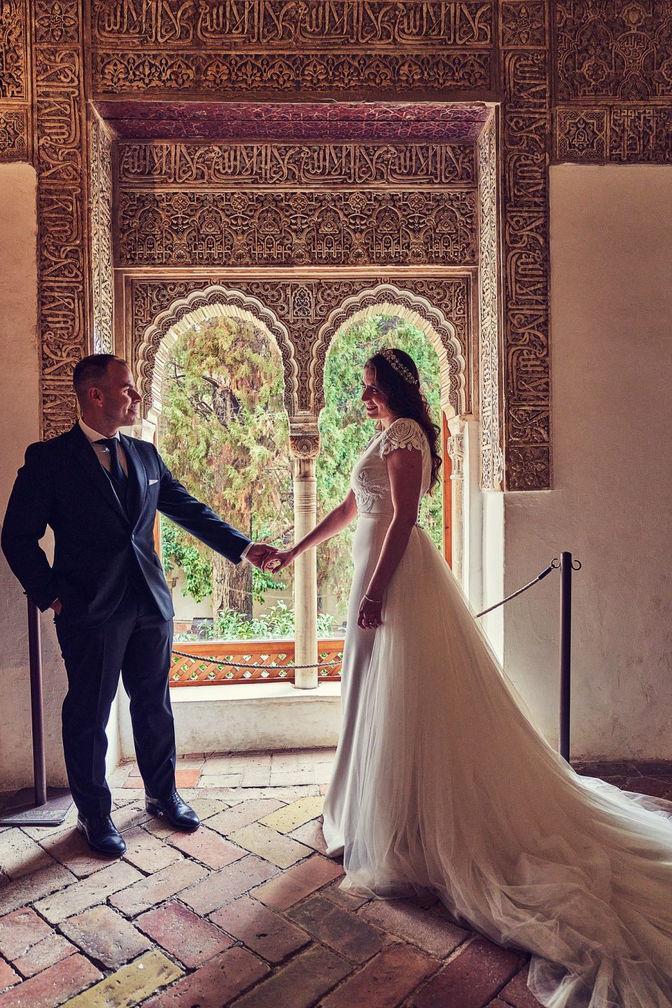Una pareja de novios se da la mano en el interior de un patio ornamentado.