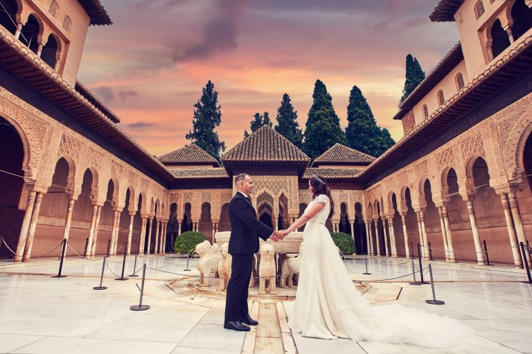 A bride and groom hold hands in the courtyard of a historic monument, surrounded by ornate arches and columns.