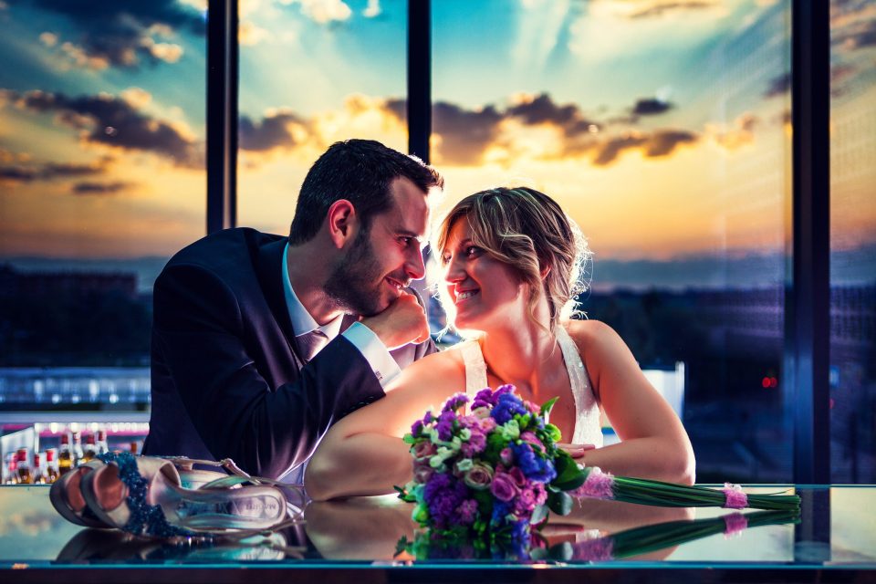 Una pareja de novios sonríe y se mira sentada junto a una mesa de cristal en el interior de un edificio.
