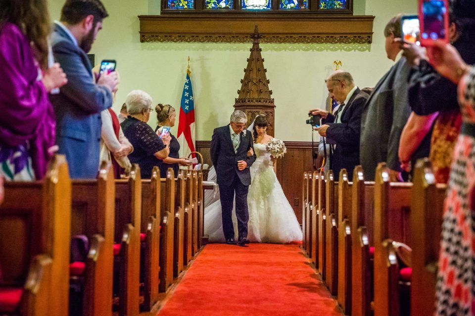A newlywed couple walks down a church aisle accompanied by an older man.