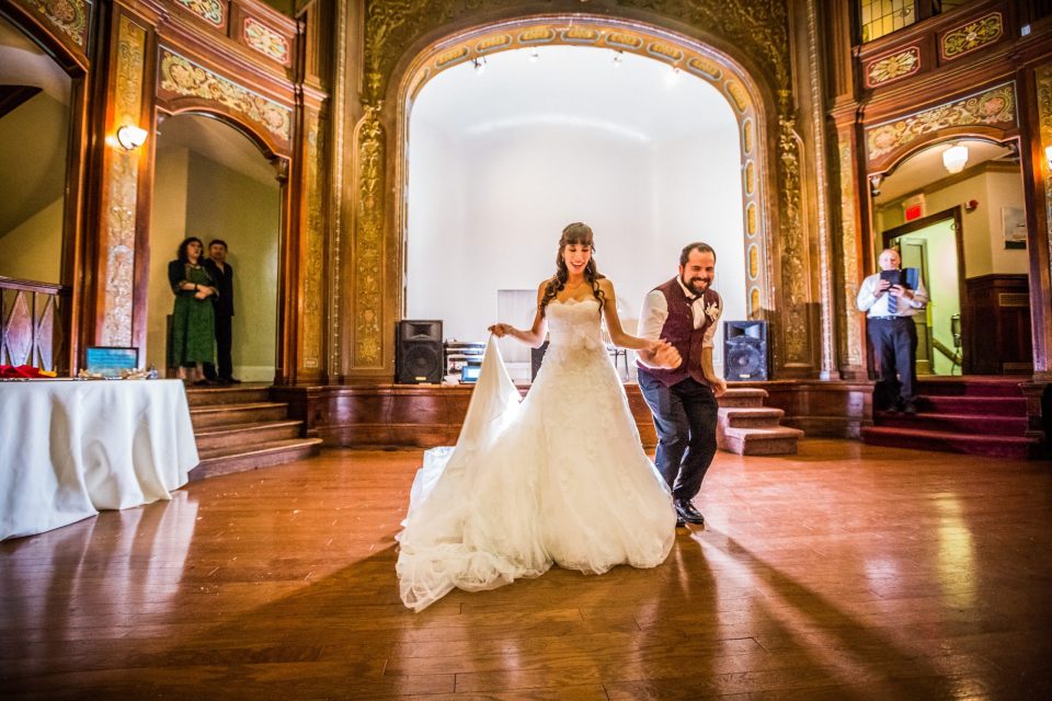 A newlywed couple dances in the centre of an ornate hall with a wooden floor.