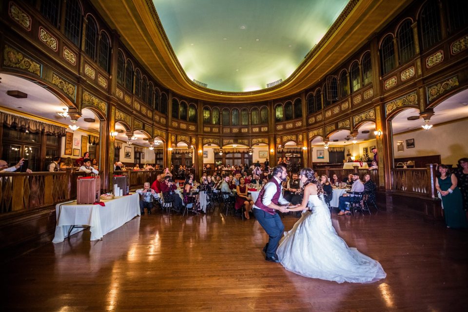 A newlywed couple dances in the foreground inside a spacious ornate hall.